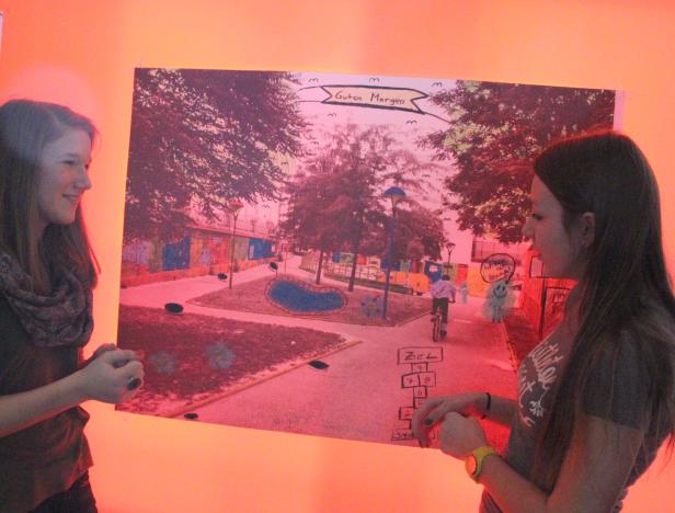 Zwei Mädchen betrachten eine Zeichnung eines Parks mit einem Radfahrer und dem Schriftzug „Guten Morgen“.