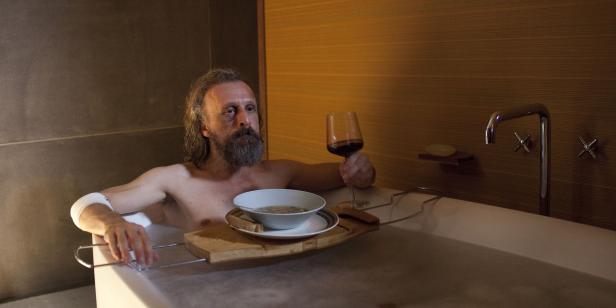 Ein Mann mit Bart entspannt mit Suppe und Wein in der Badewanne.