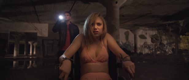 Eine gefesselte, verängstigte Frau sitzt in einem Rollstuhl, während ein Mann mit einer Taschenlampe im Hintergrund steht.