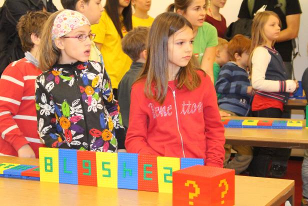Kinder stehen an einem Tisch mit bunten Bausteinen, die das Wort „BLISSHE2“ bilden.