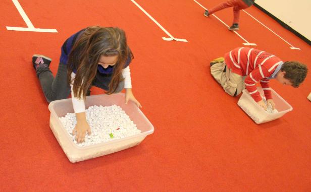 Zwei Kinder spielen mit Behältern voller Papierröllchen auf einem roten Teppich.