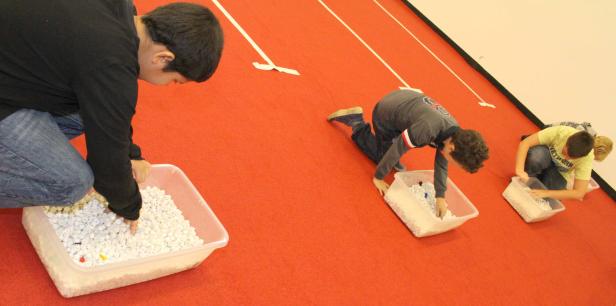 Drei Kinder suchen in Behältern mit kleinen, weißen Objekten auf einem roten Untergrund.