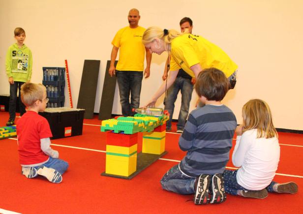 Kinder beobachten, wie eine Frau eine Wasserflasche auf eine Konstruktion aus Legosteinen stellt.