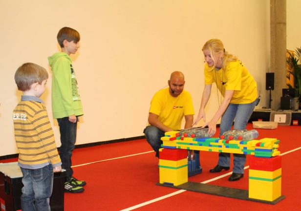 Zwei Kinder beobachten, wie eine Frau und ein Mann eine Brücke aus Legosteinen mit Wasserflaschen testen.