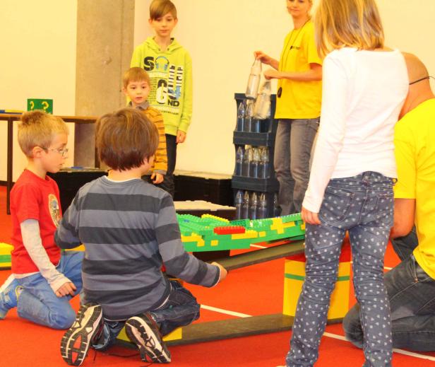 Kinder bauen gemeinsam eine Brücke aus Legosteinen, die mit Wasserflaschen belastet wird.