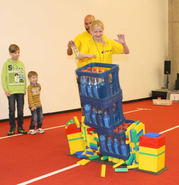 Eine Frau und ein Mann hantieren mit einem Turm aus Wasserflaschen und Legosteinen, während zwei Jungen zusehen.