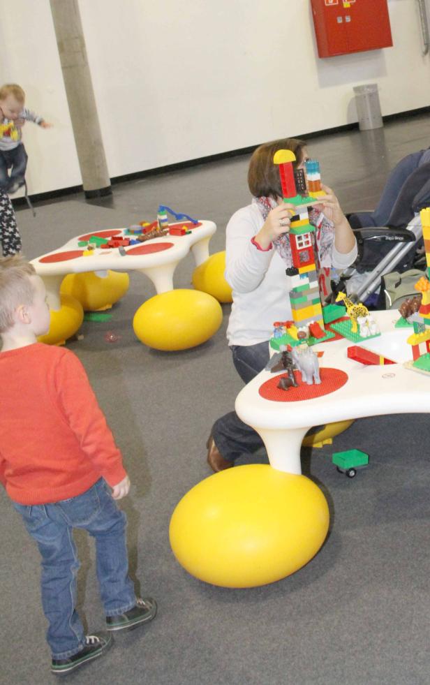 Eine Frau und Kinder spielen mit Legosteinen an einem Tisch mit gelben Sitzbällen.