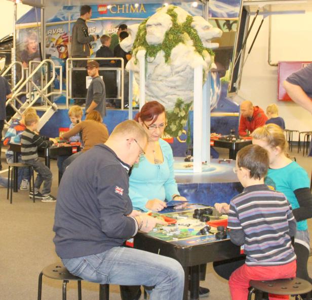 Familien bauen mit Lego-Steinen an Tischen vor einer Lego Chima-Kulisse.