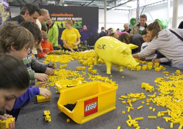 Kinder bauen gelbe Lego-Schweine an einem Tisch mit einer großen gelben Sparschwein-Statue.