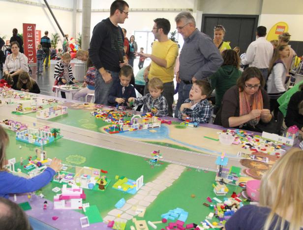 Kinder und Erwachsene bauen gemeinsam mit Legosteinen an einem großen Tisch.