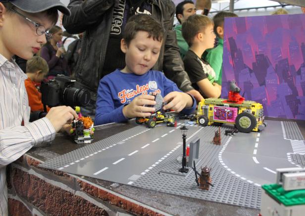 Zwei Jungen spielen mit Lego-Fahrzeugen auf einer grauen Lego-Straßenplatte.