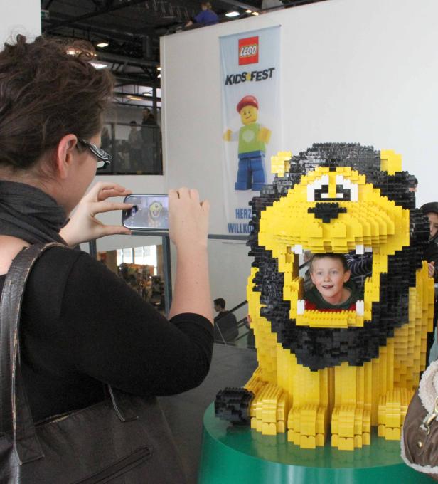 Ein Junge schaut aus dem Maul eines gelben Löwen, der aus Legosteinen gebaut wurde. Eine Frau fotografiert ihn.