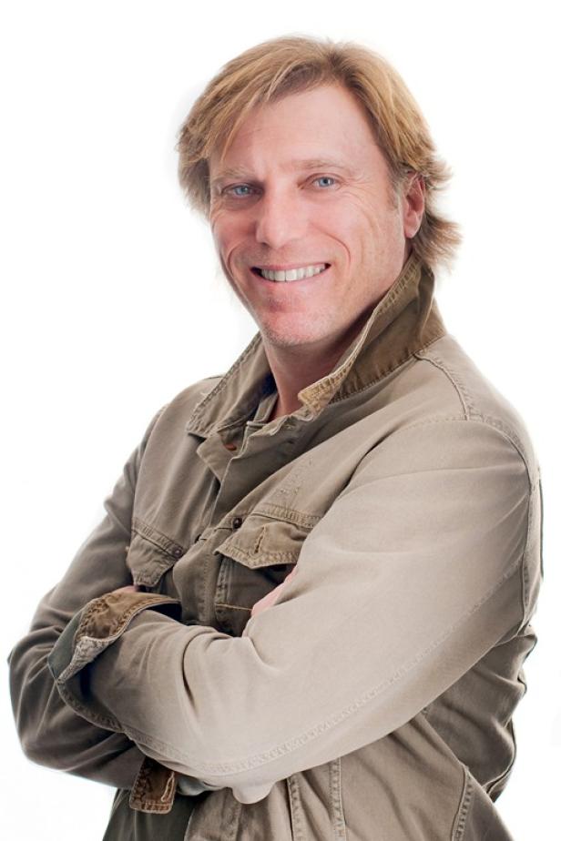 Ein lächelnder Mann mit blonden Haaren und einer olivgrünen Jacke posiert mit verschränkten Armen.