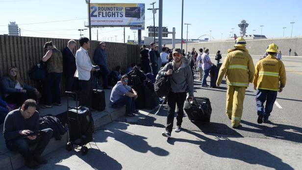 Eine Gruppe von Reisenden wartet mit Gepäck vor dem Los Angeles International Airport, während zwei Feuerwehrleute vorbeigehen.