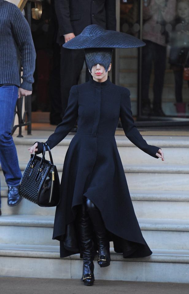 Lady Gaga in einem schwarzen Kleid mit Hut und Stiefeln auf einer Treppe.