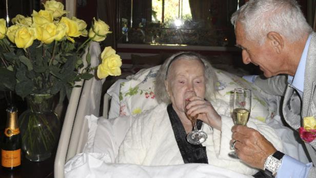 Eine ältere Frau trinkt Champagner im Bett, während ein Mann ihr zuprostet.