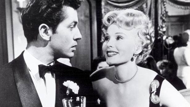 Ein Mann im Smoking und eine blonde Frau mit Halskette und Brosche schauen sich an.