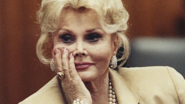 Zsa Zsa Gabor mit Schmuck und beiger Jacke, die Hand an ihr Gesicht gelegt.