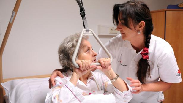 Eine ältere Frau hält sich an einem Bettgalgen fest, während eine Krankenschwester sie unterstützt.