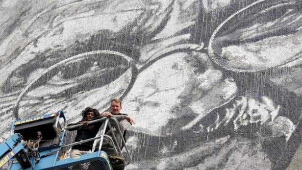 Zwei Personen auf einer Hebebühne vor einem großen Wandgemälde von John Lennon.