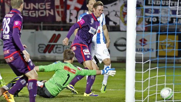 Ein Fußballspieler im lila Trikot erzielt ein Tor, während der Torwart zu spät kommt.