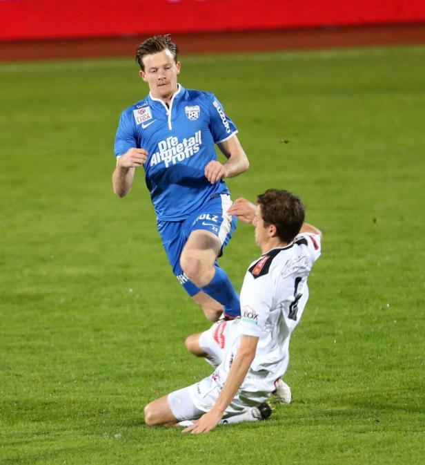 Ein Fußballspieler springt über einen am Boden liegenden Gegenspieler.