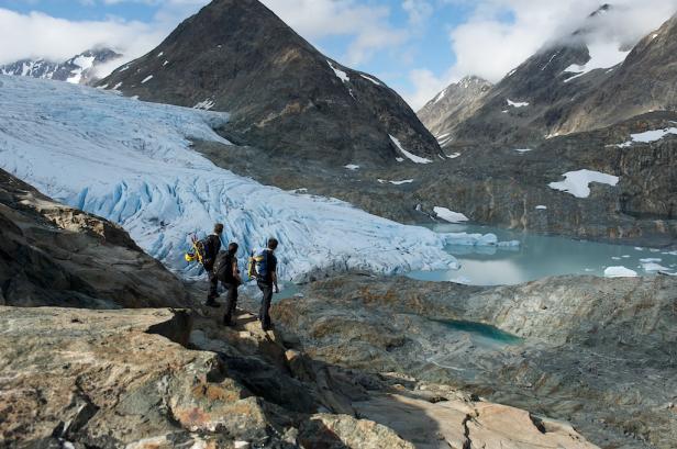 Drei Wanderer stehen auf einem Felsen mit Blick auf einen Gletscher und einen Bergsee.