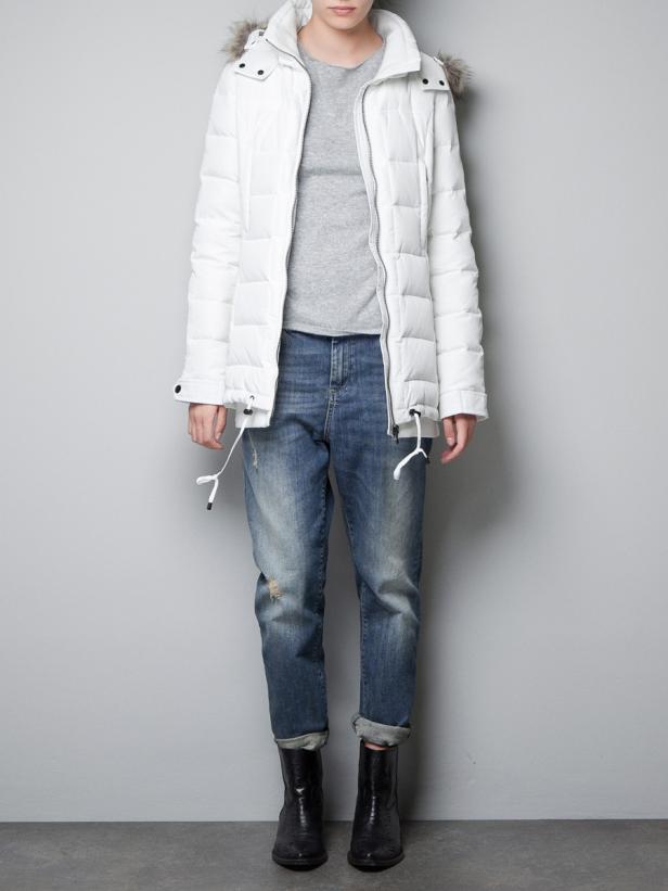 Eine Person trägt eine weiße Daunenjacke, Jeans und schwarze Stiefel vor einer grauen Wand.