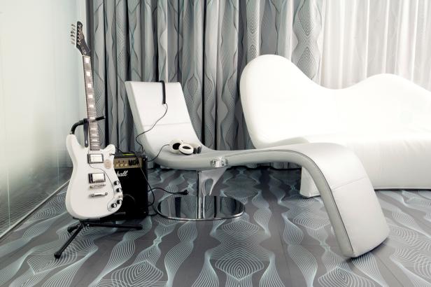 Eine weiße E-Gitarre steht neben einem Verstärker und einem modernen Sessel.