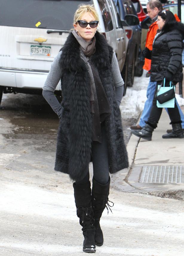 Eine Frau mit Sonnenbrille und Pelzweste geht auf einer verschneiten Straße entlang.