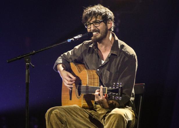 Ein bärtiger Mann mit Brille spielt Gitarre und singt ins Mikrofon.