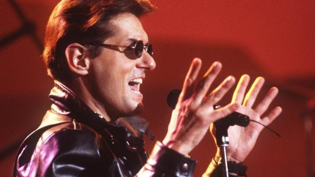 Ein Mann mit Sonnenbrille und Lederjacke singt in ein Mikrofon.