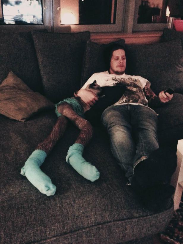 Ein Mann entspannt mit einer Katze auf der Couch und hält eine Fernbedienung in der Hand.