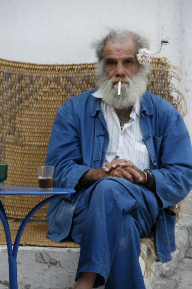 Ein älterer Mann mit Bart sitzt mit einer Zigarette im Mund in einem Sessel.