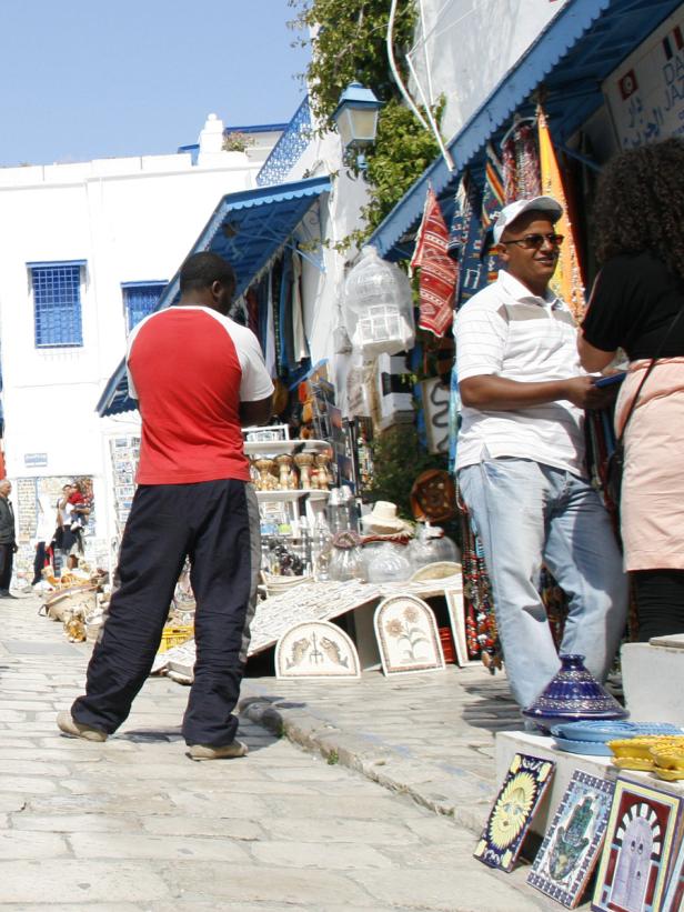 Menschen schlendern an einem sonnigen Tag durch eine Gasse mit Souvenirläden.