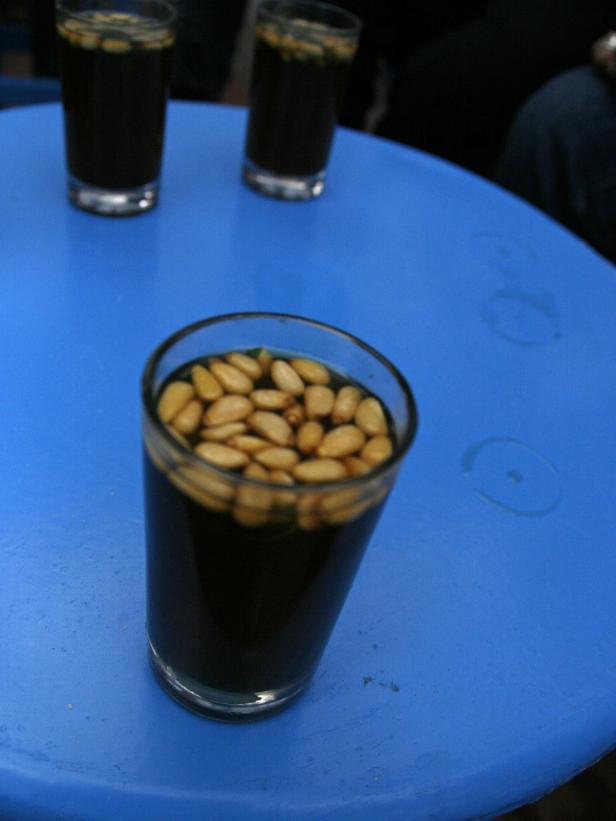 Ein Glas mit dunklem Getränk und Pinienkernen auf einem blauen Tisch.