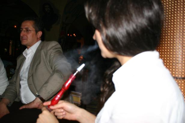 Eine Frau raucht eine Wasserpfeife, während ein Mann im Hintergrund sitzt.