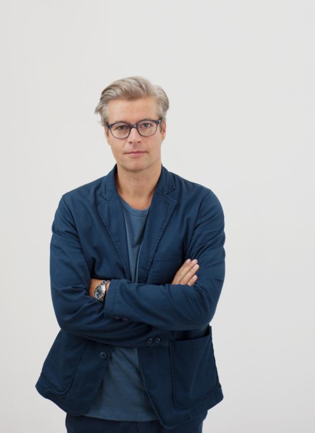 Ein Mann mit Brille und blauem Sakko steht mit verschränkten Armen vor einem weißen Hintergrund.