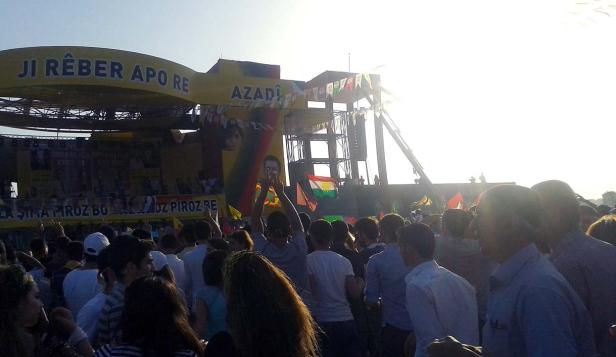 Eine Menschenmenge versammelt sich vor einer Bühne mit kurdischen Flaggen und dem Wort „Azadi“.