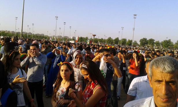 Eine große Menschenmenge versammelt sich im Freien, einige schwenken kurdische Flaggen.