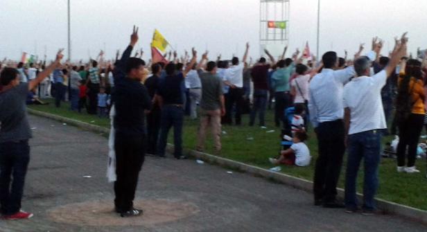 Eine Menschenmenge demonstriert mit erhobenen Händen und kurdischen Flaggen.