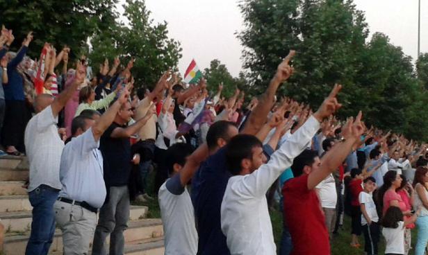 Eine Menschenmenge steht auf einer Treppe und reckt die Zeigefinger in die Luft; im Hintergrund ist eine kurdische Flagge zu sehen.