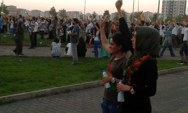Eine Menschenmenge demonstriert mit erhobenen Händen und kurdischen Flaggen im Hintergrund.