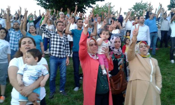 Eine Menschenmenge, darunter Frauen mit Kopftüchern und Kinder, demonstriert mit erhobenen Händen und Victory-Zeichen.