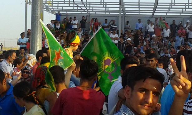 Eine Menschenmenge hält grüne Fahnen mit kurdischen Symbolen bei einer Versammlung.