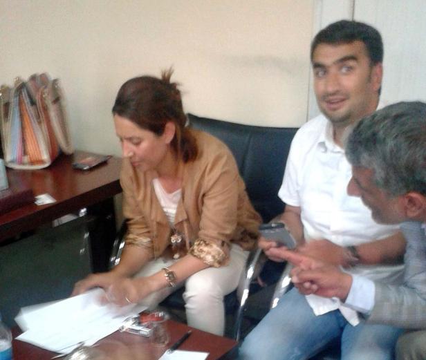 Eine Frau sitzt am Tisch und liest Papiere, während zwei Männer daneben sitzen und diskutieren.