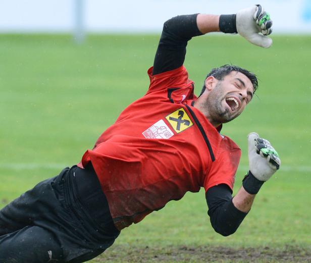Ein Torwart in rotem Trikot hechtet auf einem grünen Rasen nach dem Ball.