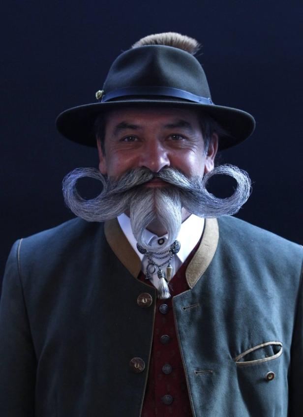 Ein Mann mit Hut und einem kunstvoll gezwirbelten Bart präsentiert sich vor dunklem Hintergrund.