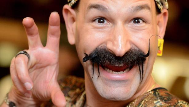 Ein Mann mit extravaganten Schnurrbart und Kopfschmuck zeigt das Victory-Zeichen.