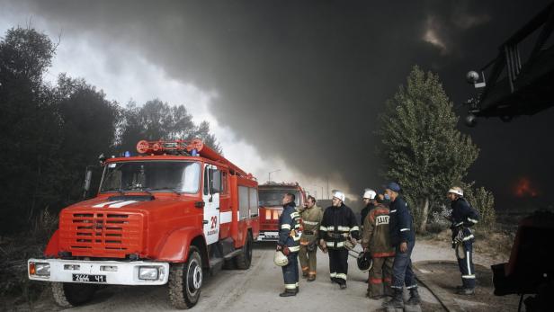 Feuerwehrmänner stehen vor roten Löschfahrzeugen, während im Hintergrund Rauch aufsteigt.